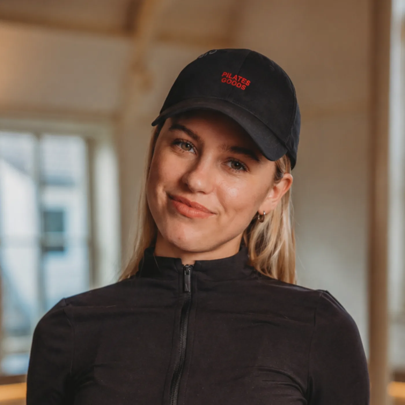 Pilates studio member wearing custom branded black dad cap with red embroidered Pilates Goods logo in bright reformer studio